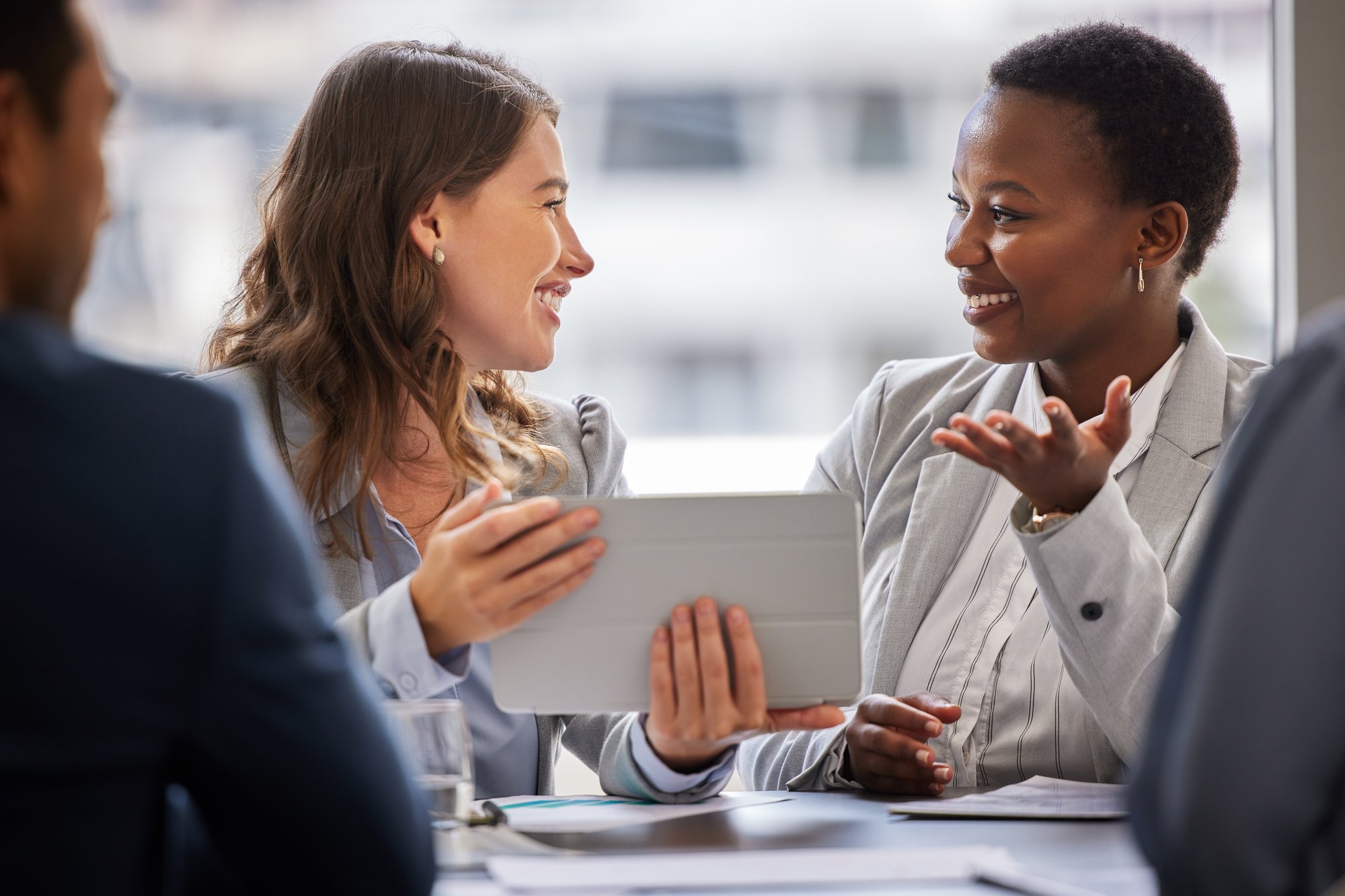 Two businesswomen smiling and discussing something on a device in a modern office setting.