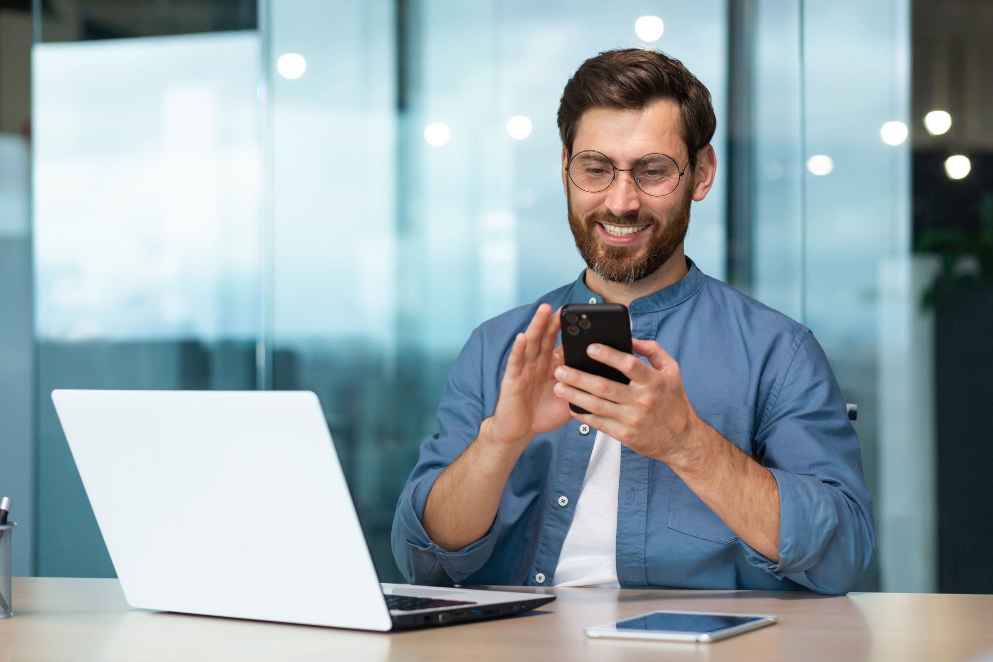 man smiling at his smartphone while sitting at a desk with a laptop and tablet in a modern office