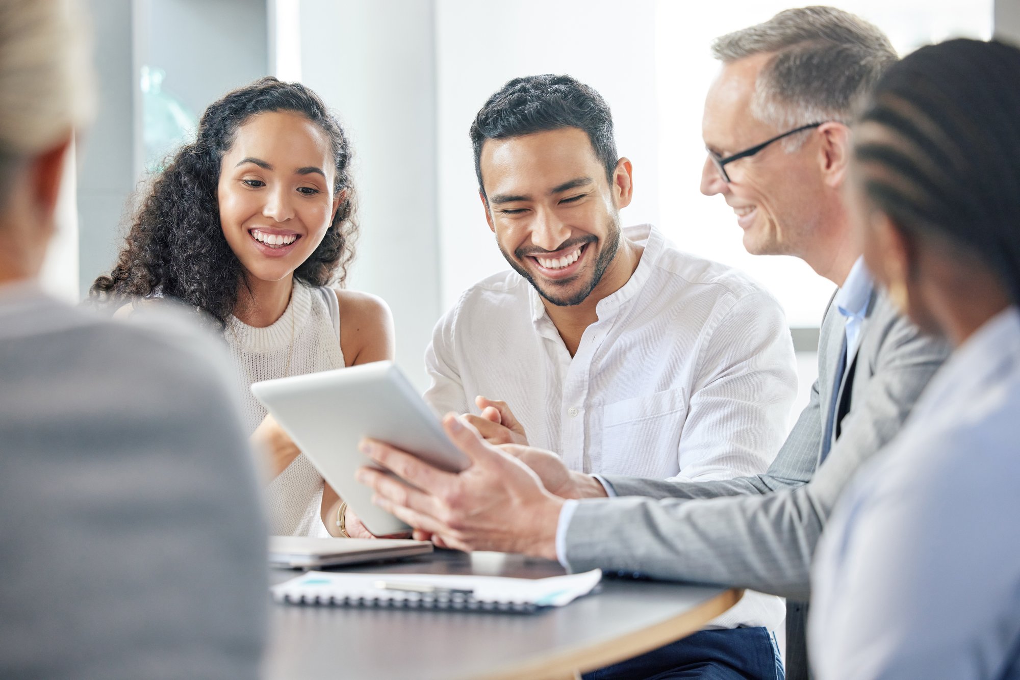 A group of diverse professionals sitting around a table, smiling and collaborating while looking at a tablet. They appear engaged and enthusiastic, suggesting a productive meeting or teamwork setting in a modern office environment.