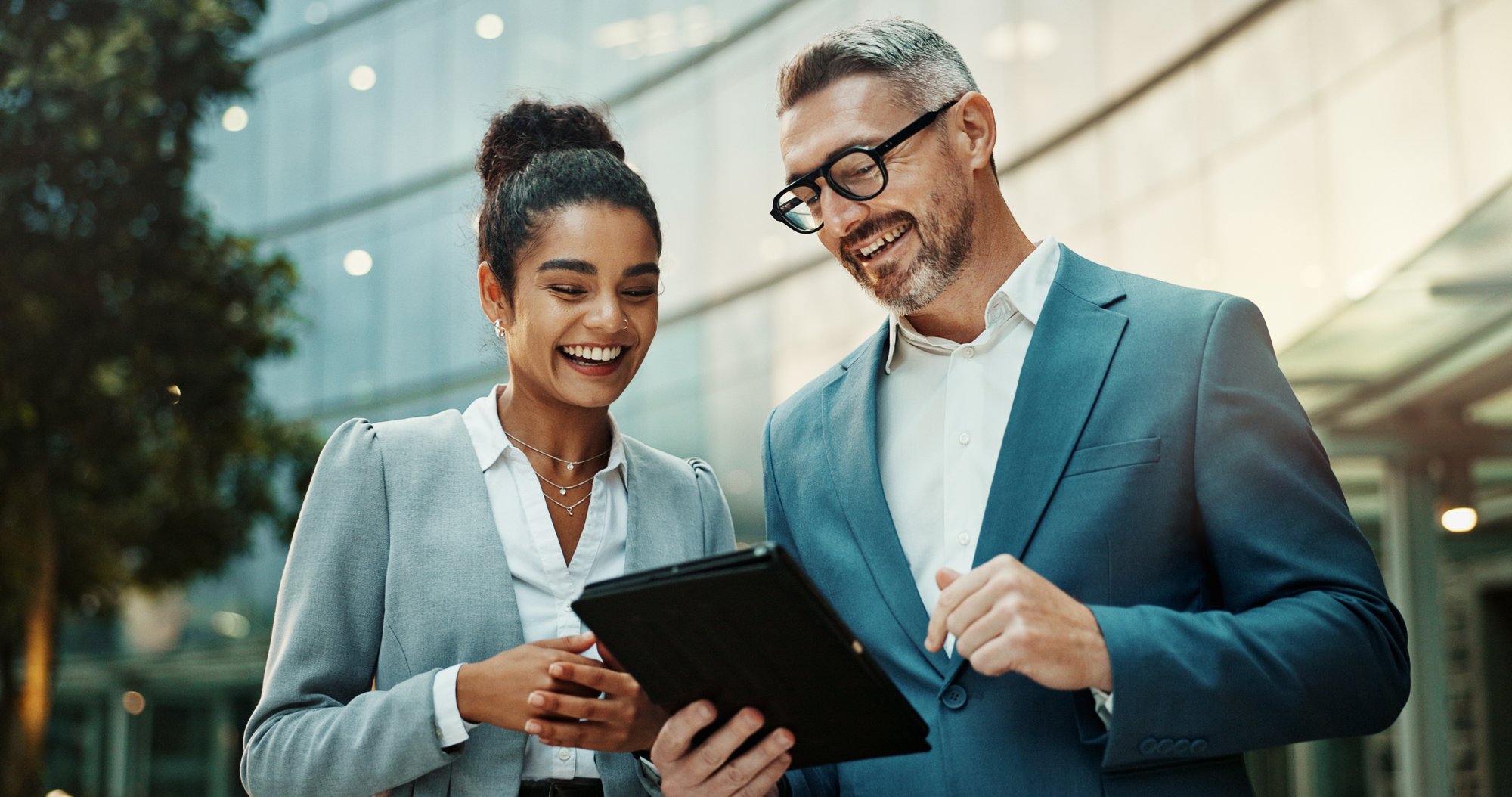 Two business professionals smiling while reviewing information on a tablet outside a modern office building, showcasing collaboration and success.